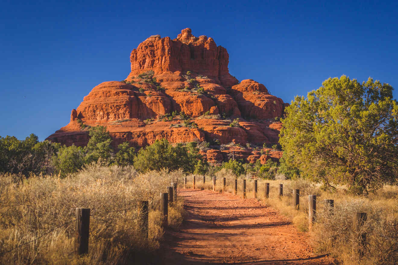 A red dirt path flanked by wooden posts leading towards a towering red rock formation under a clear blue sky in Sedona